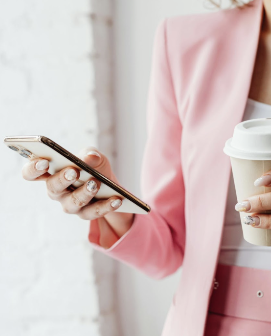 Woman in a pink blazer holding a phone and coffee, texting a real estate client as part of her marketing strategy.
